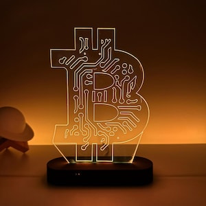 Pode incluir: Uma luz noturna de acrílico transparente em forma de símbolo Bitcoin. A luz é iluminada com um brilho branco quente, criando um visual decorativo e moderno.