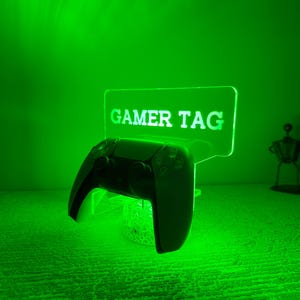 May include: A black and white gaming controller rests on a clear, illuminated stand with the words "GAMER TAG" in white. The scene is bathed in green light, with decorative figurines in the background.