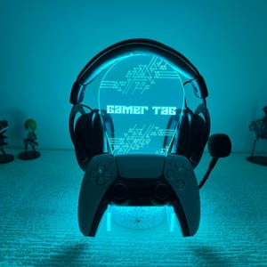 Könnte beinhalten: Gaming-LED-Lampe mit Headset und Gamecontroller. Die Lampe leuchtet blau, mit den Worten "Gamer Tag" auf der zentralen Platte. Dekorative Figuren sind im Hintergrund zu sehen.