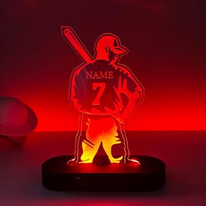 May include: A red illuminated night light featuring a baseball player silhouette holding a bat. The player is wearing a jersey with the number 7 and the word "NAME" on the back.