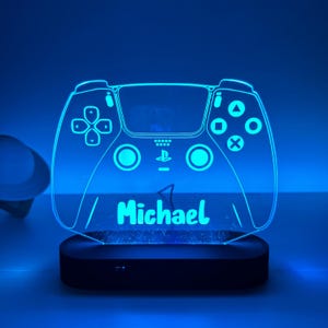 May include: A blue LED gaming controller-shaped night light. The controller design includes buttons and the name "Michael" in a glowing font. The light sits on a black base, creating a cool, modern aesthetic.