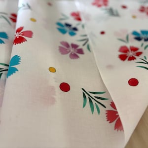 May include: White fabric with a floral pattern featuring red, blue, and purple flowers, green leaves, and yellow and red dots. The fabric is folded, showing the pattern's repeating design. The flowers have a detailed, stylized appearance.