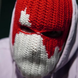 May include: A red and white crocheted balaclava. The red knit covers the head and upper face, with white accents around the eyes and mouth. The balaclava is worn over a light purple hoodie.
