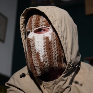 Könnte beinhalten: Eine Person trägt eine beige Kapuzenjacke und eine braun, weiß und beige gestreifte Häkel-Balaclava. Die Sturmhaube bedeckt Kopf und Hals, mit Augenöffnungen. Die Jacke hat eine Kapuze mit Kordelzug und ein kleines Logo auf der Brust.
