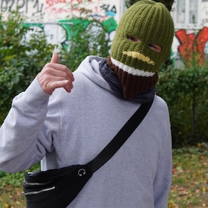 Könnte beinhalten: Eine Person trägt eine grüne Strick-Balaclava mit gelben, weißen und braunen Streifen, einen grauen Kapuzenpullover und eine schwarze Bauchtasche. Die Bauchtasche hat einen silbernen Reißverschluss und einen schwarzen Riemen.