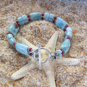 May include: A blue and red gemstone bracelet with a silver toggle clasp. The bracelet is made with rectangular and round beads. The bracelet is photographed on a white starfish on a sandy background.