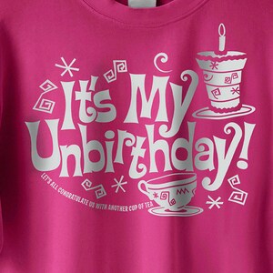 Op de afbeelding: Roze T-shirt met een wit grafisch ontwerp met de tekst "It's My Unbirthday!" met een theekopje en taartontwerp. De tekst onder de grafiek luidt "Laten we allemaal op onszelf toasten met nog een kopje thee."
