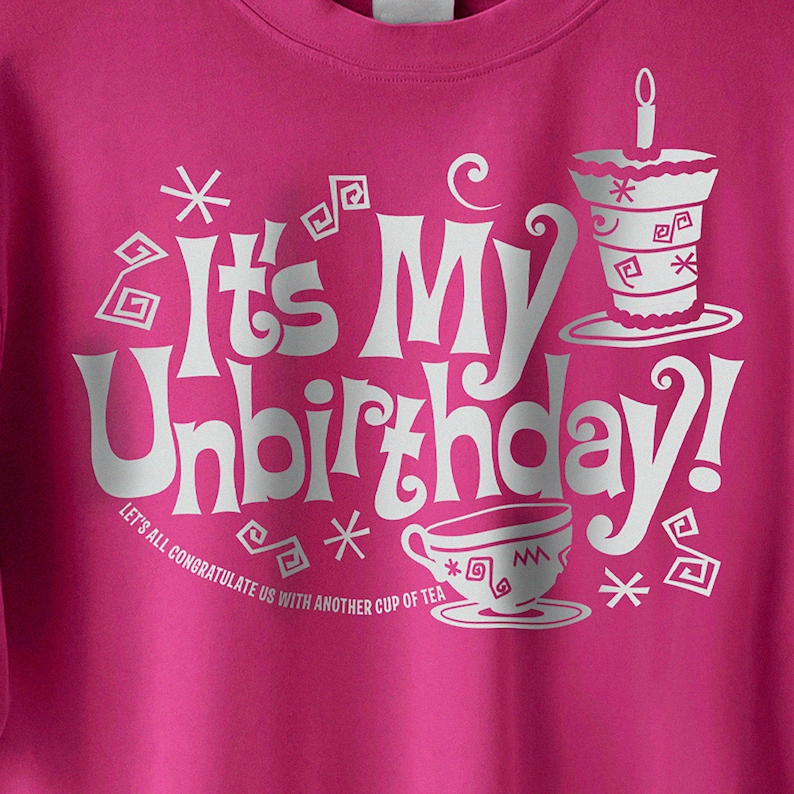Op de afbeelding: Roze T-shirt met een wit grafisch ontwerp met de tekst "It's My Unbirthday!" met een theekopje en taartontwerp. De tekst onder de grafiek luidt "Laten we allemaal op onszelf toasten met nog een kopje thee."