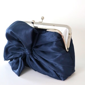 May include: A navy blue satin clutch bag with a large bow on the front. The bag has a silver clasp.