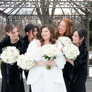 May include: A group of five individuals posing together, with one person in a white strapless gown and white faux fur wrap, surrounded by four others in black floor-length dresses with black faux fur wraps, all holding white rose bouquets.