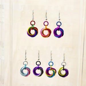May include: A selection of chainmaille earrings in various colours. Each earring comprises interlocking metal rings, creating a circular design. The earrings are displayed on a clear background, highlighting the different colour combinations and styles.