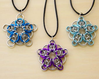 Puede incluir: Tres colgantes de flores hechos a mano con cota de malla. Cada colgante presenta un diseño de flor de cinco pétalos con un anillo central. Los colgantes son de color azul, morado y azul claro, con anillos plateados. Cuelgan de cordones negros.
