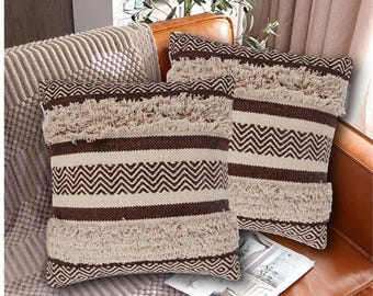 May include: Two brown and cream striped throw pillows with fringe trim. The pillows have a chevron pattern and are arranged on a brown leather couch.