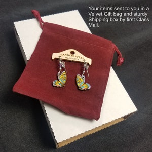 K&ouml;nnte beinhalten: Schmetterlingsohrringe mit gelben, blauen und rosa Fl&uuml;geln, pr&auml;sentiert auf einem Holzanh&auml;nger mit der Aufschrift "HANDCRAFTERS Made in USA". Die Ohrringe befinden sich in einem bordeauxroten Samtgeschenkbeutel, auf einer wei&szlig;en Pappschachtel.