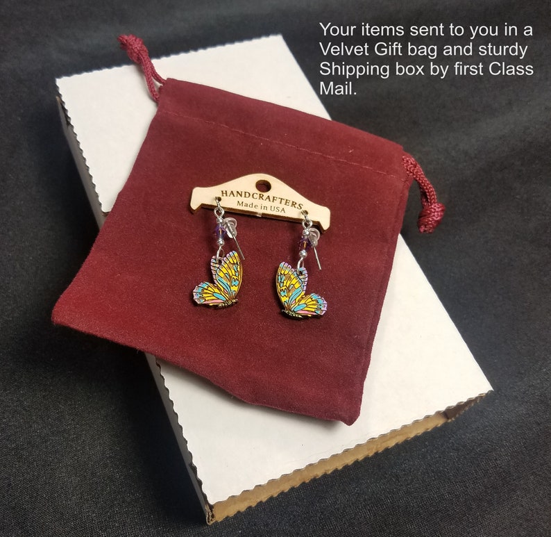 K&ouml;nnte beinhalten: Schmetterlingsohrringe mit gelben, blauen und rosa Fl&uuml;geln, pr&auml;sentiert auf einem Holzanh&auml;nger mit der Aufschrift "HANDCRAFTERS Made in USA". Die Ohrringe befinden sich in einem bordeauxroten Samtgeschenkbeutel, auf einer wei&szlig;en Pappschachtel.