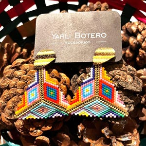 May include: A pair of colourful geometric triangle earrings with gold accents. The earrings are made of small beads and have a geometric pattern. The earrings are on a brown background.