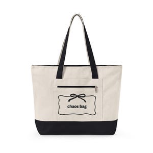 May include: A cream-coloured canvas tote bag with black handles and a black base. The front features a zipped pocket with a black graphic of a rectangular frame, a bow, and the text "chaos bag" printed in the centre in a simple black font.