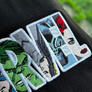 May include: Black embroidered patch with the word "MARVEL" in white outlined letters. The letters are filled with images of Marvel characters, including Hulk, Thor, and Black Widow.