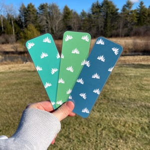 Pu&ograve; includere: Tre segnalibri trasparenti con un motivo di castello bianco su sfondo turchese, verde e blu. I segnalibri sono tenuti in una mano su uno sfondo di prato verde.