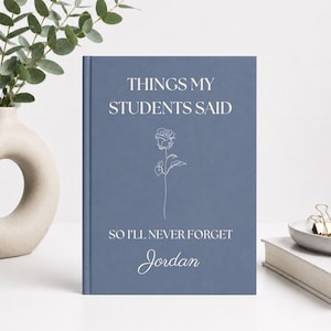 May include: A dusty blue hardcover journal featuring the white text "THINGS MY STUDENTS SAID SO I'LL NEVER FORGET" and the name "Jordan" in a script font. A simple white line-art rose illustration is centered between the text lines.