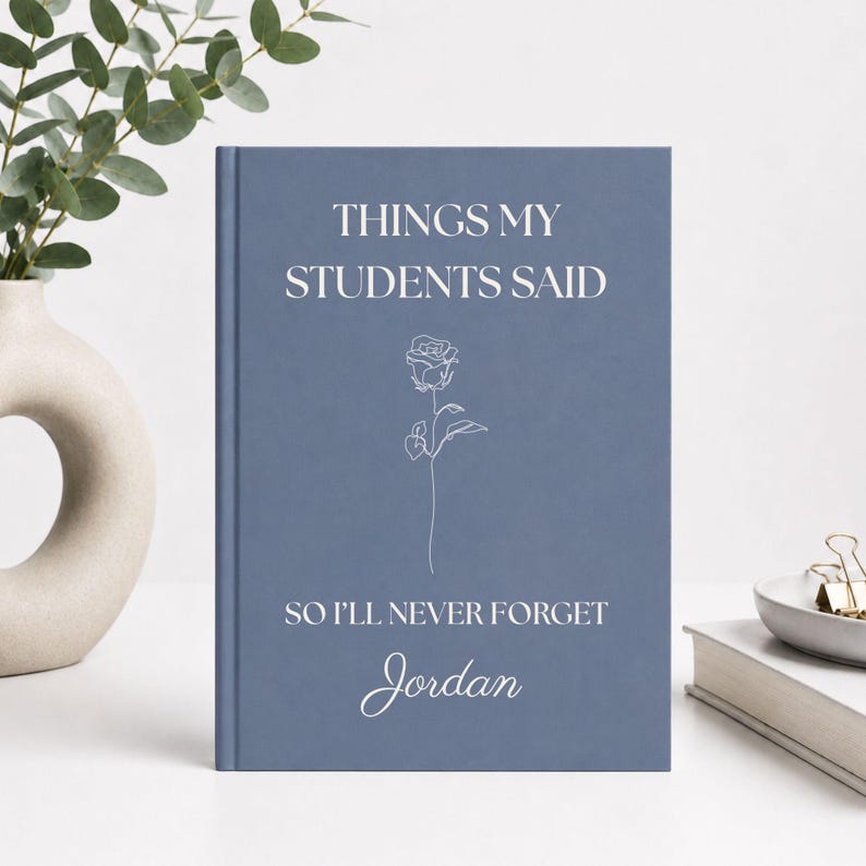 May include: A dusty blue hardcover journal featuring the white text "THINGS MY STUDENTS SAID SO I'LL NEVER FORGET" and the name "Jordan" in a script font. A simple white line-art rose illustration is centered between the text lines.