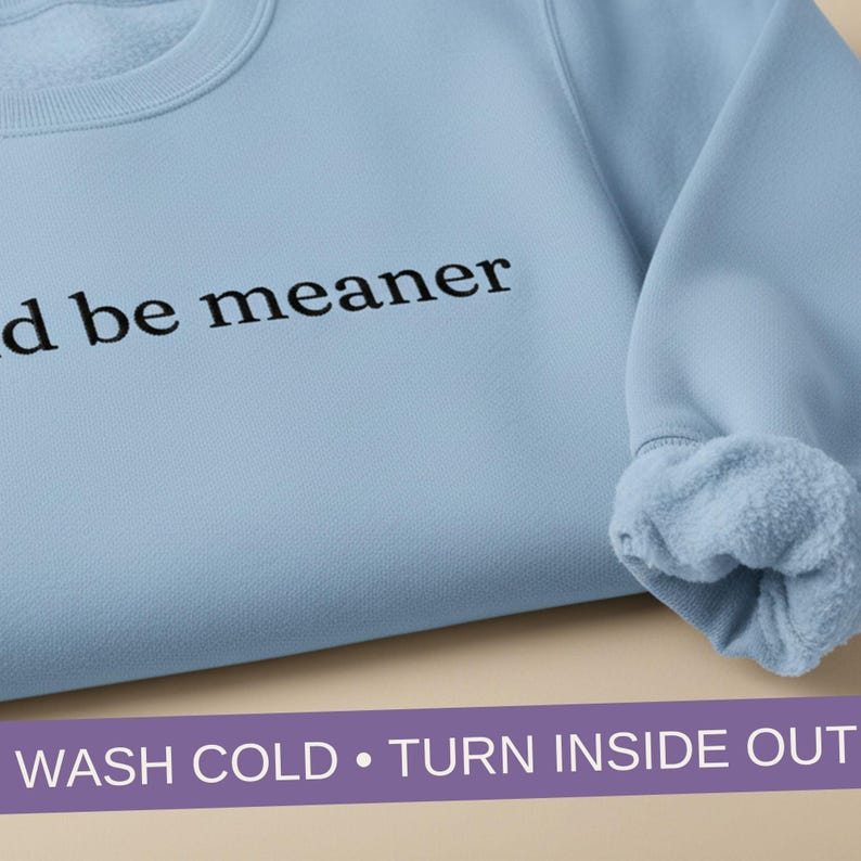May include: A pale blue sweatshirt with the words "d be meaner" printed in black. The sleeve cuff reveals a soft, fleecy interior. The bottom of the image displays the text "WASH COLD &bull; TURN INSIDE OUT".