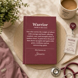 Op de afbeelding: Een bordeauxrood hardcover boek met de definitie van het woord Warrior, dat iemand beschrijft die de last van anderen met moed en hart draagt, inclusief de naam Jessica onderaan. Het boek ligt op een ronde, gevlochten onderzetter.