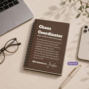 May include: A dark brown spiral-bound notebook with white text that reads Chaos Coordinator, a definition of the term, and the name Jordan. The notebook is positioned next to a laptop, a smartphone, and a pen on a light-coloured surface.