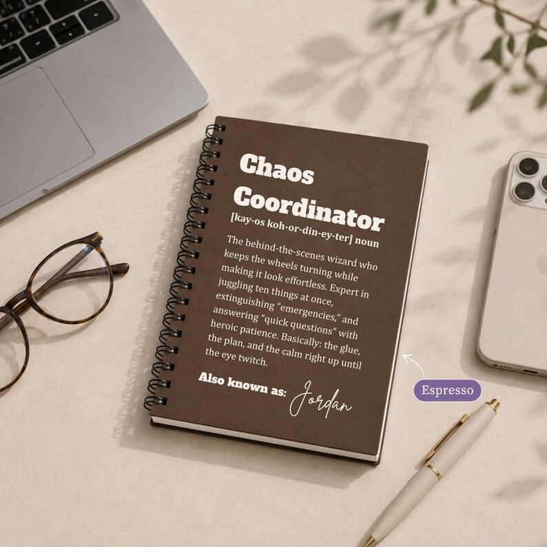 May include: A dark brown spiral-bound notebook with white text that reads Chaos Coordinator, a definition of the term, and the name Jordan. The notebook is positioned next to a laptop, a smartphone, and a pen on a light-coloured surface.