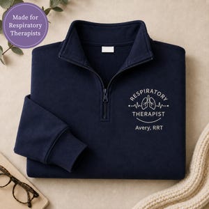 May include: A navy blue quarter-zip pullover featuring an embroidered white graphic of lungs with a heartbeat line and the text "RESPIRATORY THERAPIST" above the name "Avery, RRT". The garment has a high collar and a central zipper closure.