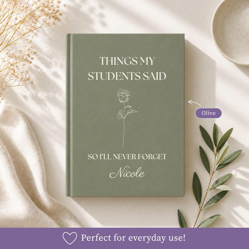 May include: An olive green hardcover journal with the text "THINGS MY STUDENTS SAID SO I'LL NEVER FORGET" and the name "Nicole" printed in white. A thin, white line-art illustration of a single rose is centered between the lines of text.