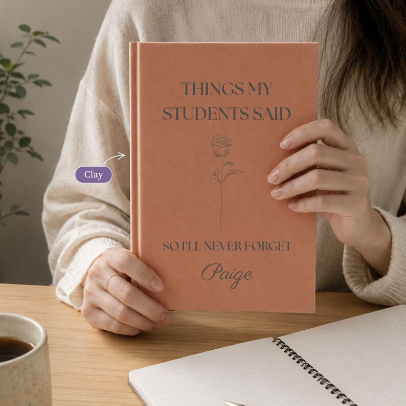 May include: A clay-colored hardcover journal with the text "THINGS MY STUDENTS SAID SO I'LL NEVER FORGET" and the name "Paige" printed in dark gray. A thin, line-art illustration of a single rose is centered between the lines of text.