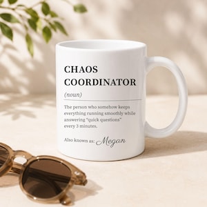 Może przedstawiać: Biały ceramiczny kubek z czarnym nadrukiem o treści CHAOS COORDINATOR (noun). Tekst zawiera definicję dotyczącą utrzymywania wszystkiego w płynnym działaniu oraz frazę Also known as: Megan zapisaną kursywną czcionką.