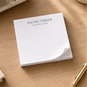 May include: A square white notepad with a stack of paper sheets. The top sheet features the printed black text RACHEL PARKER and FINANCIAL ANALYST in a serif font. The bottom right corner of the top sheet is slightly curled upward.