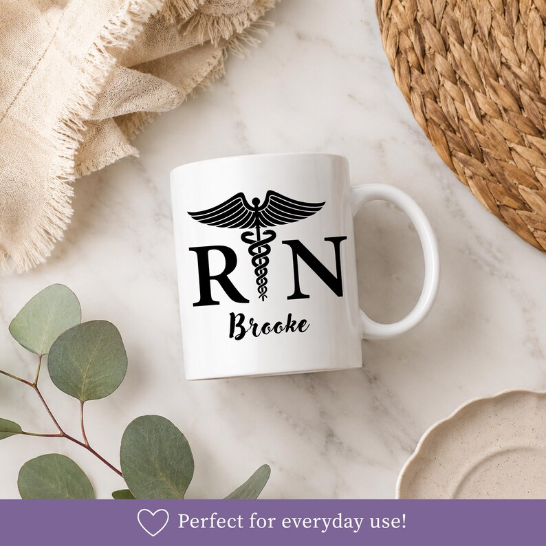 May include: A white ceramic mug featuring a black caduceus symbol positioned between the letters R and N, with the name Brooke printed in a black script font below the letters. The mug has a standard handle and a smooth, glossy finish.