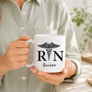 May include: A white ceramic mug featuring a black graphic of a caduceus symbol positioned between the letters R and N. Below the symbol, the name Lauren is printed in a black script font. The mug has a smooth finish and a curved handle.