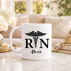 May include: A white ceramic mug featuring a black caduceus symbol centered between the letters R and N. Below the symbol, the name Alexis is printed in a black script font. The mug has a smooth finish and a curved handle on the left side.