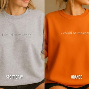 May include: Two sweatshirts, one sport gray and one orange, each with the phrase "i could be meaner" printed in a simple font across the chest. The sport gray sweatshirt has "SPORT GRAY" printed below the text, and the orange one has "ORANGE" printed below.