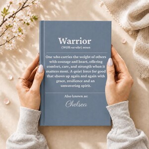 Op de afbeelding: Een leigrijs hardcover boek met de definitie van het woord Warrior, gevolgd door de tekst Also known as: Chelsea. De kaft bevat witte gedrukte tekst die de definitie van een krijger omschrijft als iemand die het gewicht van anderen draagt.