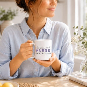 Puede incluir: Una taza de cer&aacute;mica blanca con el texto Registered NURSE en una combinaci&oacute;n de letra cursiva negra y letras may&uacute;sculas moradas, seguido del nombre EMILY y el texto EST. 2026 en letras may&uacute;sculas negras centrado en la parte frontal.
