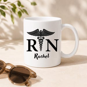 May include: A white ceramic mug featuring a black graphic of a caduceus symbol centered between the letters R and N. Below the symbol, the name Rachel is printed in a black script font. The mug has a standard handle and a smooth finish.