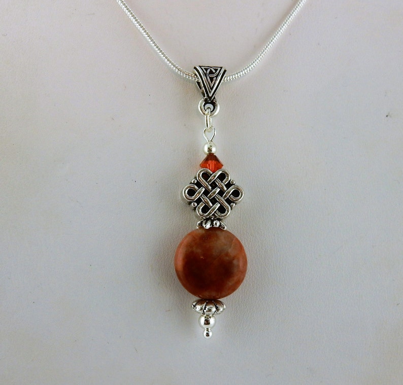 May include: A silver chain necklace with a silver Celtic knot pendant and a brown stone bead. The pendant has a small red crystal bead at the top.