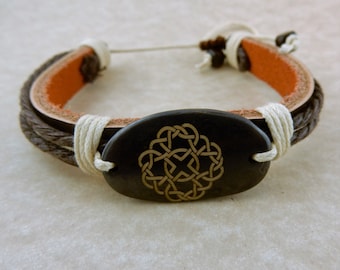 May include: A brown leather bracelet with a white cord and a black oval charm featuring a gold Celtic knot design.
