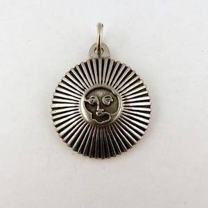 May include: Silver pendant with a sun design featuring a face in the center. The sun has a textured, radiating design.