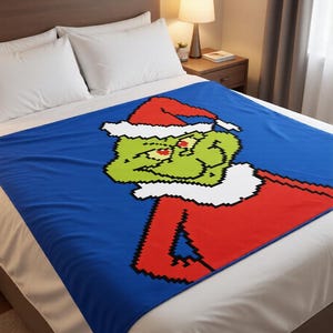May include: A rectangular throw blanket featuring a pixelated graphic of a green character wearing a red Santa hat and a red coat with white trim. The design is set against a solid blue background, showcasing a blocky, digital art style.