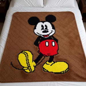 May include: A brown blanket featuring a pixelated image of Mickey Mouse. The blanket has a diagonal textured pattern. Mickey Mouse is depicted in his classic outfit with black ears, a white face, red shorts, and yellow shoes.