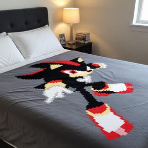 May include: A gray blanket featuring a pixelated image of a black, red, and white character. The character is in a running pose, with red and white accents on the feet. The blanket is on a bed with white pillows and a black headboard.