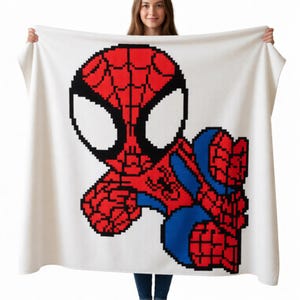 May include: A white rectangular throw blanket featuring a large, centered pixel art graphic of a superhero character. The character is rendered in red, blue, and black, showcasing a stylized mask with large white eyes and a web-like pattern.