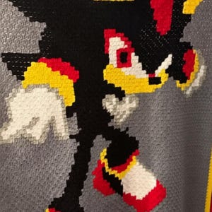 May include: A crochet blanket featuring a pixelated graphic of the character Shadow the Hedgehog in black, red, white, and yellow yarn. The character is set against a textured gray background with a yellow and red border along the bottom edge.
