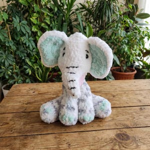 May include: A plush white elephant with light blue inner ears and accents. The elephant has a pink neck and gray and white patterned legs and feet. The toy is sitting on a wooden surface with a green leafy background.
